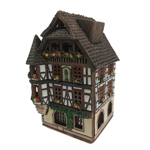 May include: A brown and white ceramic model of a traditional German half-timbered house with a steeply pitched roof, decorated with red and yellow flowers and green vines.
