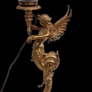 May include: A gold-colored, ornate table lamp featuring a winged figure holding a torch, topped with a frosted glass shade. The lamp has a detailed base and stem, with a decorative design. The lamp is a decorative lighting fixture.