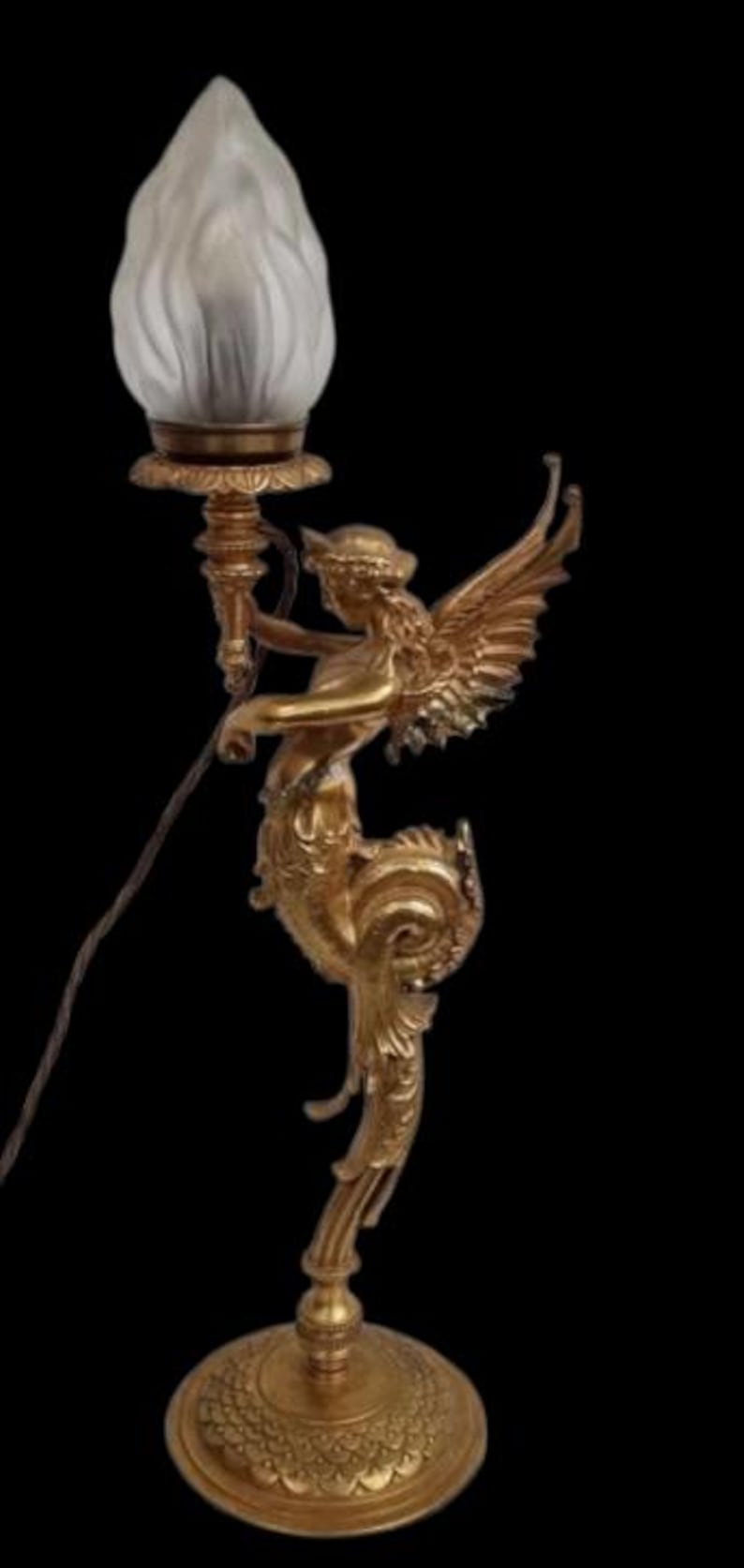 May include: A gold-colored, ornate table lamp featuring a winged figure holding a torch, topped with a frosted glass shade. The lamp has a detailed base and stem, with a decorative design. The lamp is a decorative lighting fixture.