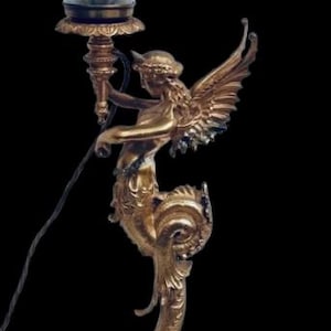 May include: Ornate gold-colored table lamp featuring a winged figure holding a torch, topped with a frosted glass shade. The lamp has a detailed base and a decorative, flowing design. The lamp is approximately 18 inches tall.
