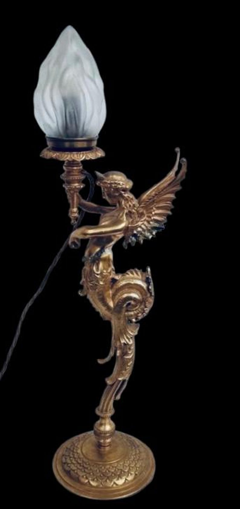 May include: Ornate gold-colored table lamp featuring a winged figure holding a torch, topped with a frosted glass shade. The lamp has a detailed base and a decorative, flowing design. The lamp is approximately 18 inches tall.