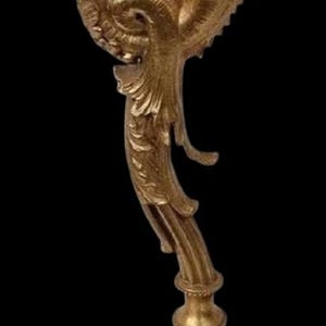 May include: Ornate gold-colored decorative object with a detailed, swirling design. The object has a base with a textured pattern and a curved, stylized top. The background is black, highlighting the object's intricate details.