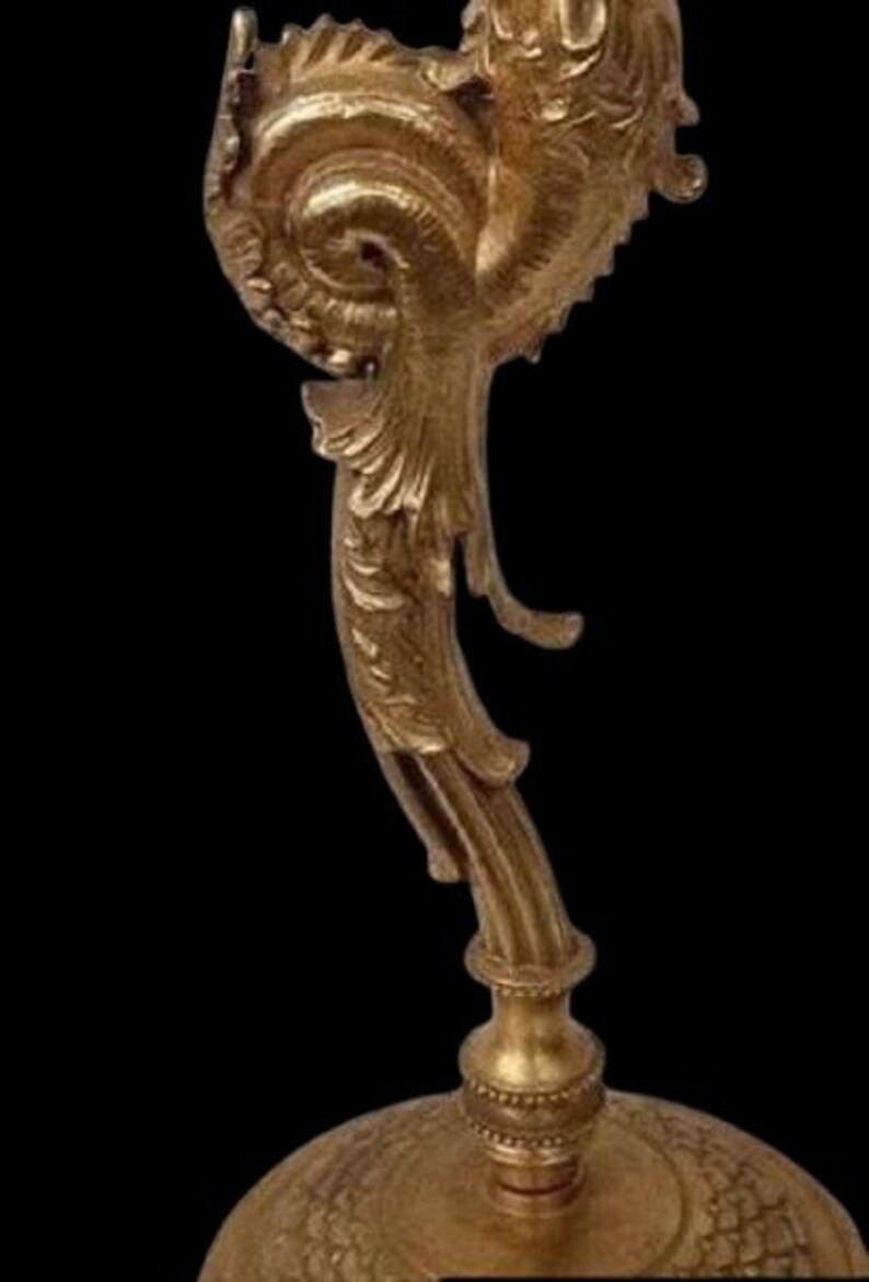 May include: Ornate gold-colored decorative object with a detailed, swirling design. The object has a base with a textured pattern and a curved, stylized top. The background is black, highlighting the object's intricate details.