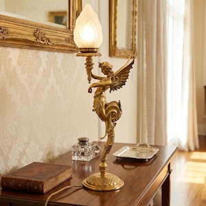 May include: A gold-toned table lamp featuring a winged figure holding a torch-shaped light fixture. The lamp has a detailed, ornate design and sits on a dark wooden table. A book and inkwell are also on the table.