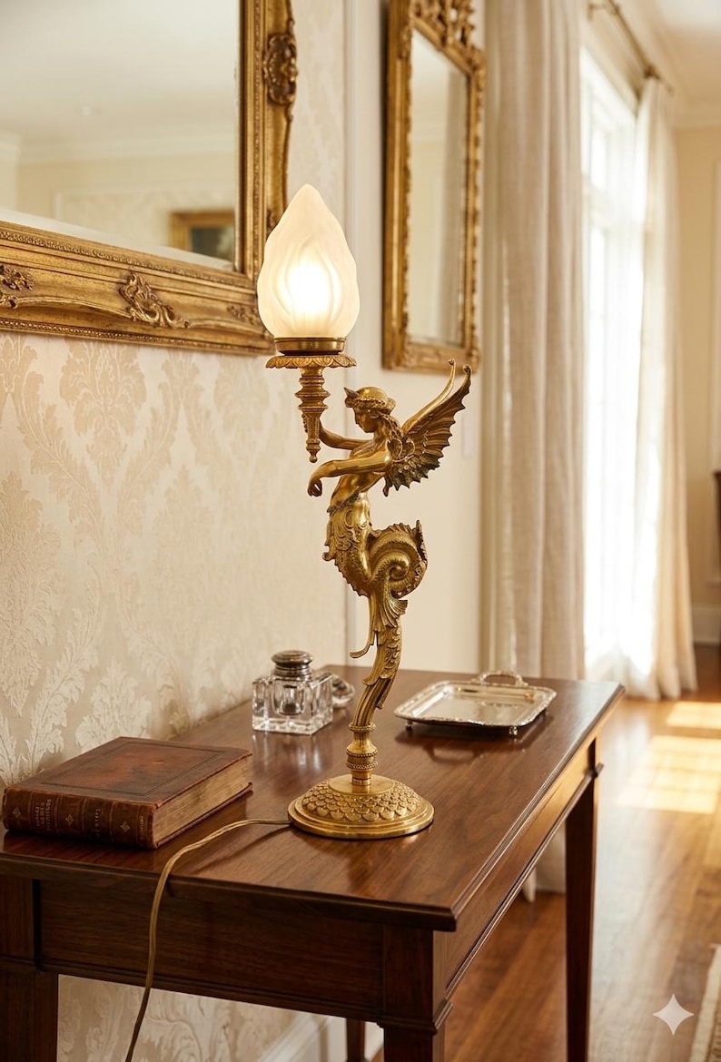 May include: A gold-toned table lamp featuring a winged figure holding a torch-shaped light fixture. The lamp has a detailed, ornate design and sits on a dark wooden table. A book and inkwell are also on the table.