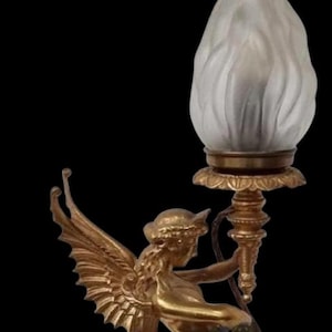 May include: A gold-toned lamp featuring a winged figure holding a torch-shaped light fixture. The light has a frosted, flame-like appearance. The lamp has an ornate base and is set against a black background.
