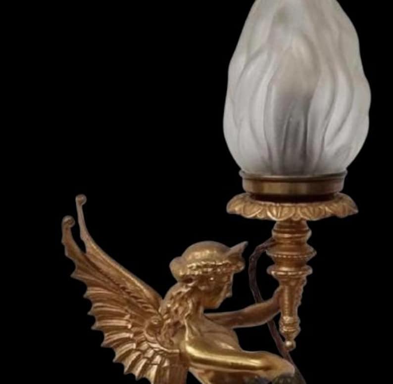 May include: A gold-toned lamp featuring a winged figure holding a torch-shaped light fixture. The light has a frosted, flame-like appearance. The lamp has an ornate base and is set against a black background.