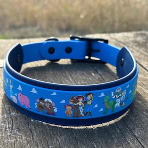 May include: A bright blue pet collar featuring a repeating pattern of Toy Story characters including Woody, Buzz Lightyear, Jessie, Bullseye, Mr. and Mrs. Potato Head, Rex, and Hamm. The collar has a black buckle and hardware with a dark blue inner lining.