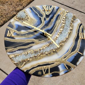 May include: A black, white, and gold resin clock with Roman numeral markings and a gold glitter finish. The clock face features a white and black agate design with clear crystals embedded in the resin.