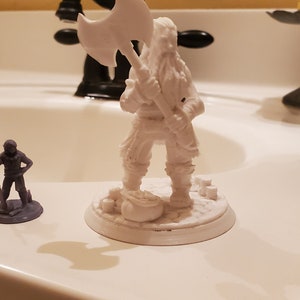 May include: A white plastic miniature figurine of a bearded warrior holding a large axe. The figurine is standing on a round base with a small bag of coins at its feet. A smaller gray plastic miniature figurine of a warrior holding a staff is also visible in the image.