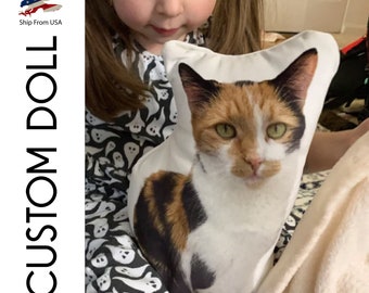 Può includere: Un cuscino bianco con un'immagine stampata di un gatto calico. Il gatto sta guardando direttamente la telecamera. Il cuscino è tenuto da una bambina che indossa un vestito bianco e nero con un motivo a fantasmi.