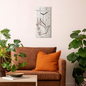 Puede incluir: Un reloj de pared rectangular con una superficie de espejo. El reloj presenta un diseño minimalista con manecillas y marcadores de hora negros. Un contorno marrón de una paloma que lleva una rama de olivo está debajo de la esfera del reloj. El reloj está montado en una pared blanca.