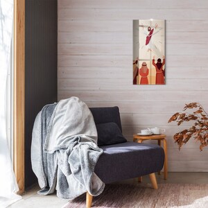 Puede incluir: Una obra de arte religiosa enmarcada representa una figura con una túnica roja ascendiendo, rodeada de luz, con figuras debajo. La obra de arte se exhibe en una pared de madera blanca, encima de un sillón gris oscuro con una manta gris.