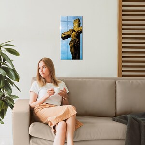 May include: A woman sits on a beige couch, holding a white mug, looking to the left. A framed print of a golden crucifix against a blue sky hangs on the wall behind her.