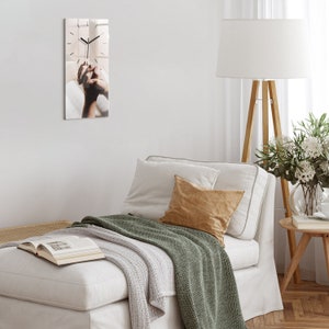 Puede incluir: Un reloj de pared rectangular con un diseño de veta de madera clara. Las manecillas del reloj son negras y la esfera del reloj presenta una imagen de manos. El reloj está montado en una pared blanca, con un sofá blanco y almohadas en primer plano.
