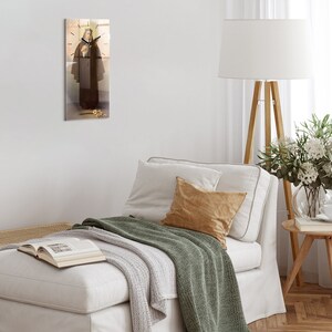 Puede incluir: Un reloj de pared de vidrio con una esfera blanca y agujas negras. El reloj presenta una pintura de una mujer con una túnica marrón que sostiene un libro. El reloj está colgado en una pared blanca.