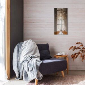 Puede incluir: Un reloj de pared rectangular con una imagen de catedral cuelga de una pared de madera blanca. Un sillón gris oscuro con una manta y un cojín grises está frente a la pared. Una pequeña mesa auxiliar de madera tiene un juego de té.