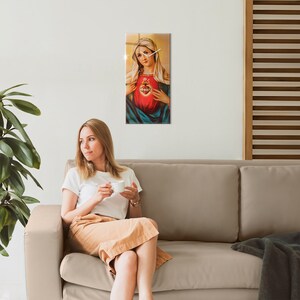 Puede incluir: Una mujer se sienta en un sofá beige, sosteniendo una taza blanca. Detrás de ella, en la pared, hay un reloj con una imagen religiosa de la Virgen María con un corazón.