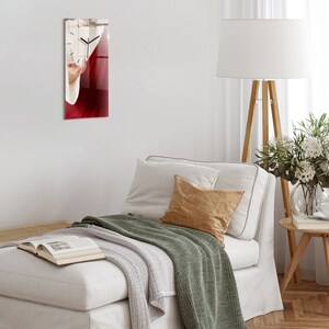 Puede incluir: Un reloj de pared de vidrio con fondo rojo y esfera blanca. Las manecillas del reloj apuntan a las 12:00. El reloj está colgado en una pared blanca.