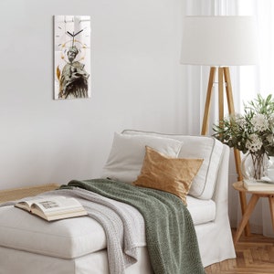 Puede incluir: Un sofá blanco con una manta de tiro verde y una almohada de color canela. Un reloj con un marco dorado y un fondo blanco está colgado en la pared sobre el sofá. El reloj presenta una imagen en blanco y negro de un hombre con un halo y un bastón.