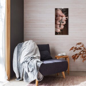 Puede incluir: Un sillón gris con una manta gris drapeada sobre él. Una pequeña mesa auxiliar de madera con una taza y un platillo blancos se encuentra junto al sillón. Una pared marrón y blanca con un reloj que representa una mano sosteniendo un rosario y un crucifijo cuelga de la pared.