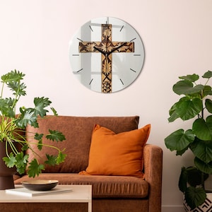 May include: A round glass clock with a brown wooden cross design. The clock is hanging on a white wall above a brown leather sofa with an orange cushion.