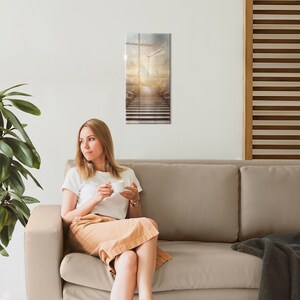 May include: A woman sits on a beige couch, holding a white mug, looking to the side. A clock with a white face and black hands is hanging on the wall behind her. The clock face is a surreal image of a stairway leading up to a bright light.