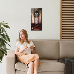 Puede incluir: Una mujer se sienta en un sofá beige, sosteniendo una taza blanca, mirando hacia la izquierda. Un reloj con una imagen del interior de una iglesia está colgado en la pared detrás de ella.