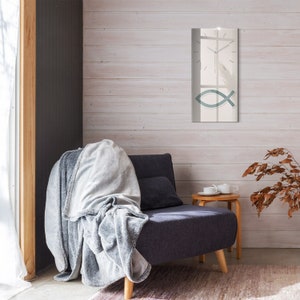 Puede incluir: Una manta gris drapeada sobre un sillón gris oscuro con marco de madera. Una pared blanca con un espejo rectangular que presenta un símbolo de pez estilizado y una esfera de reloj se encuentra detrás del sillón. Una pequeña mesa de madera con una taza de té blanca y un platillo se encuentra a la derecha del sillón.