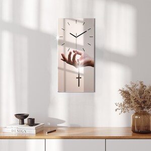 Puede incluir: Un reloj con una cruz colgando de la aguja de los segundos. El reloj está colocado sobre un fondo rosa claro con una mano que se extiende para sostener la cruz.