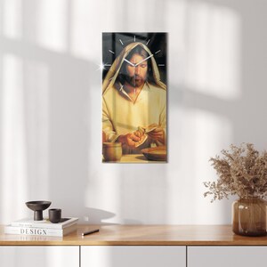 Puede incluir: Un reloj con fondo negro y agujas blancas. La esfera del reloj es una pintura de Jesús partiendo el pan. El reloj está colgado en una pared blanca.