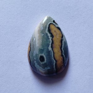 May include: A teardrop-shaped gemstone with a polished surface. The stone features a mix of colors, including tan, gray, and white, with dark brown and black accents. The gemstone has a unique pattern and is likely used in jewelry.