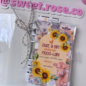 May include: A clear, spiral-bound savings challenge notebook with a cow and sunflower design. The cover reads "Just a girl saving her MOOO-Lah!" with the text "savings challenge" and "Save $900". A pearl beaded chain is attached.