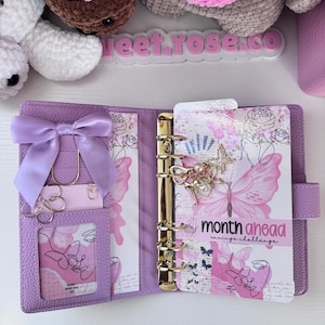 May include: Open purple planner with gold rings, a bow, and butterfly-themed inserts. The planner features a pocket with a card and gold paper clips. The text "month ahead savings challenge" is visible on one of the inserts.