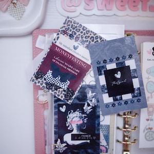 Puede incluir: Una agenda de anillas rosa que contiene tarjetas decorativas con varios diseños, incluyendo una tarjeta con estampado de leopardo con el texto "MONEYFESTING", una tarjeta de cuadros azules con el texto "thatgirl" y una tarjeta con una estatua y "I'M RICH".