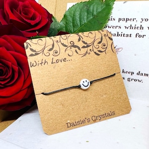 May include: A black string bracelet with a white smiley face charm. The bracelet is on a brown card with the text "With Love..." and "Daisie's Crystals".