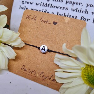 May include: A black string bracelet with a silver charm featuring the letter "A". The bracelet is displayed on a brown card with the text "With love..." and "Daisie's Crystals" on it.