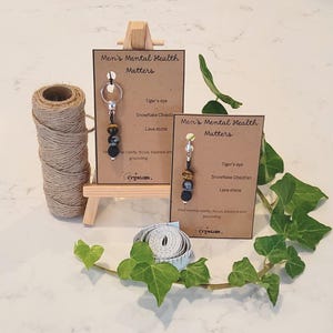 May include: Two brown cards with the text "Men's Mental Health Matters" on them. Each card has a small keychain with black, brown, and gray beads attached. The text on the cards also lists the types of stones used in the keychains: Tiger's eye, Snowflake Obsidian, and Lava stone.