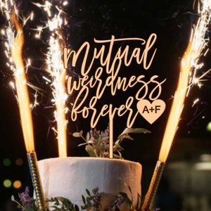 Op de afbeelding: Een witte bruidstaart met een houten taarttopper waarop "Mutual weirdness forever" staat met een hartje waarop "A+F" staat. De taart is versierd met groen en er zijn vonken aan weerszijden van de taarttopper.