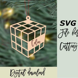 Può includere: Un ornamento a forma di cubo in legno con un design a griglia, personalizzato con il nome "Colin". L'ornamento è appeso a un filo rosso su uno sfondo verde sfocato. Il testo a destra recita "File SVG per il taglio."