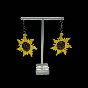 Puede incluir: Un par de pendientes de sol amarillos con un centro de purpurina plateada. Los pendientes están colgados de un soporte blanco.
