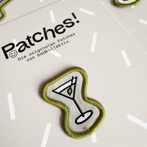 Pu&ograve; includere: Toppa ricamata raffigurante un bicchiere da martini con un'oliva, bordata con filo verde oliva su uno sfondo grigio chiaro. La toppa &egrave; presentata su un cartoncino con il testo Patches! Die originalen Patches von badkollektiv.