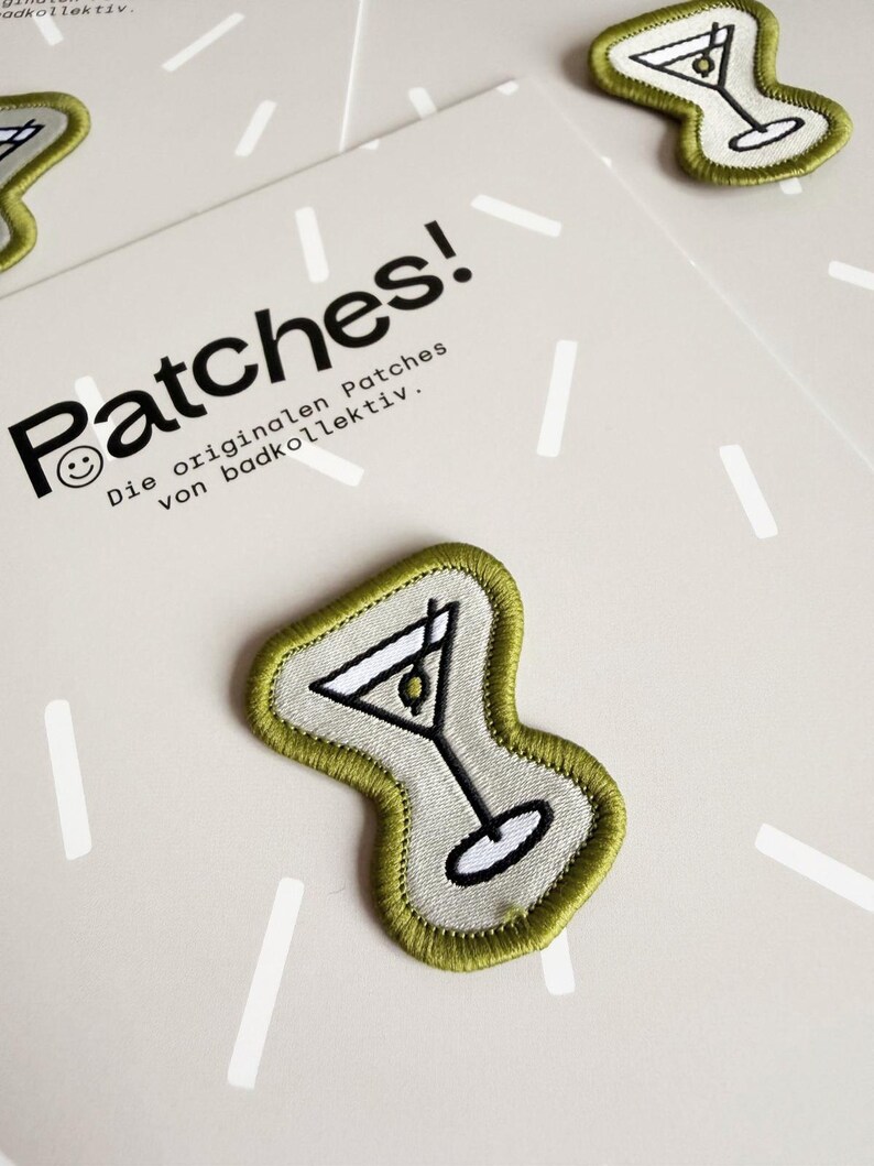 Pu&ograve; includere: Toppa ricamata raffigurante un bicchiere da martini con un'oliva, bordata con filo verde oliva su uno sfondo grigio chiaro. La toppa &egrave; presentata su un cartoncino con il testo Patches! Die originalen Patches von badkollektiv.