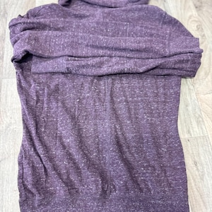 K&ouml;nnte beinhalten: Ein lila Kapuzenpullover mit Kordelzugkapuze und Vordertasche. Der Kapuzenpullover ist aus einem weichen, melierten Stoff gefertigt. Das Kleidungsst&uuml;ck ist auf einer Holzoberfl&auml;che gefaltet.