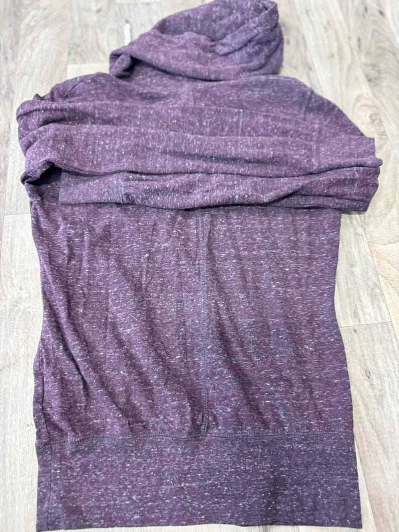 K&ouml;nnte beinhalten: Ein lila Kapuzenpullover mit Kordelzugkapuze und Vordertasche. Der Kapuzenpullover ist aus einem weichen, melierten Stoff gefertigt. Das Kleidungsst&uuml;ck ist auf einer Holzoberfl&auml;che gefaltet.