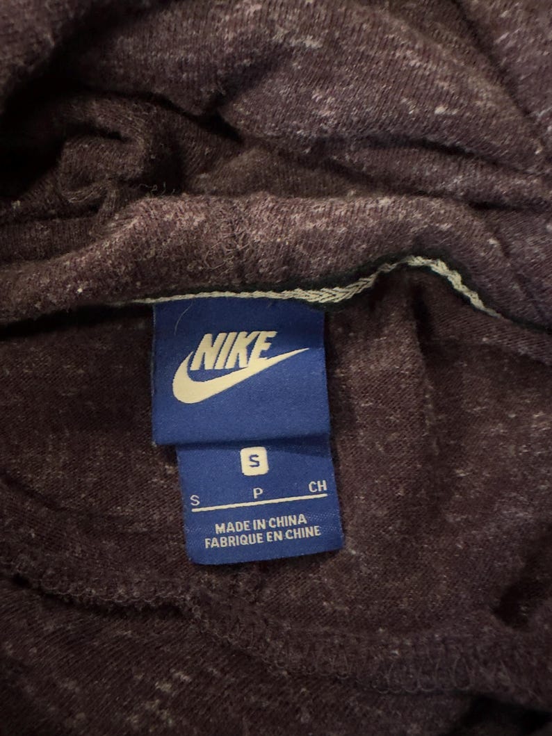 K&ouml;nnte beinhalten: Nahaufnahme eines dunkelvioletten Hoodies mit einem kleinen wei&szlig;en Fleckenmuster. Ein blaues Etikett mit dem Nike-Logo und dem Text "S P CH" ist sichtbar. Auf dem Etikett steht auch "MADE IN CHINA" und "FABRIQU&Eacute; EN CHINE".