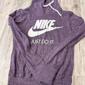K&ouml;nnte beinhalten: Ein lila Nike Hoodie mit dem Markennamen und "JUST DO IT" in wei&szlig;en Buchstaben. Der Hoodie hat einen Kordelzug und lange &Auml;rmel mit gerippten B&uuml;ndchen. Der Stoff hat eine gesprenkelte Textur.