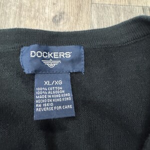 May include: Close-up of a black sweater with a navy blue tag. The tag reads "DOCKERS" and includes the size "XL/XG". The tag also states the sweater is 100% cotton and made in Hong Kong. The care instructions are "REVERSE FOR CARE."