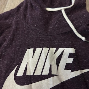 K&ouml;nnte beinhalten: Dunkelvioletter Nike-Hoodie mit wei&szlig;em Logo und Kordelzug. Der Hoodie hat eine gesprenkelte Textur und einen hohen Ausschnitt. Das Nike-Logo ist gro&szlig; und mittig auf der Vorderseite.