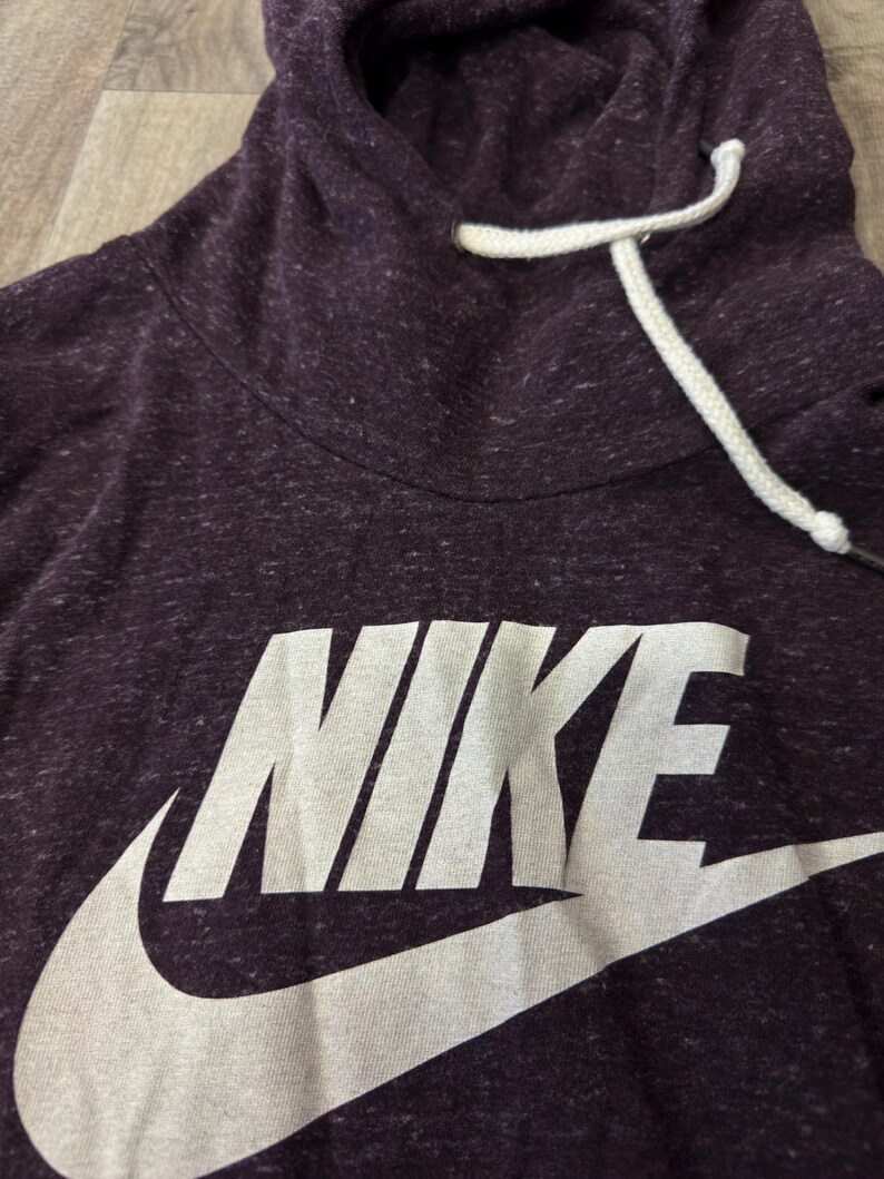 K&ouml;nnte beinhalten: Dunkelvioletter Nike-Hoodie mit wei&szlig;em Logo und Kordelzug. Der Hoodie hat eine gesprenkelte Textur und einen hohen Ausschnitt. Das Nike-Logo ist gro&szlig; und mittig auf der Vorderseite.